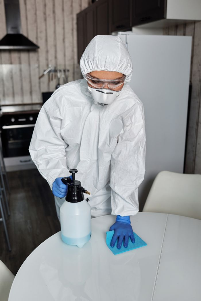 who-we-are Une personne entièrement équipée en EPI désinfectant une table à l’intérieur pour l’hygiène et la sécurité.