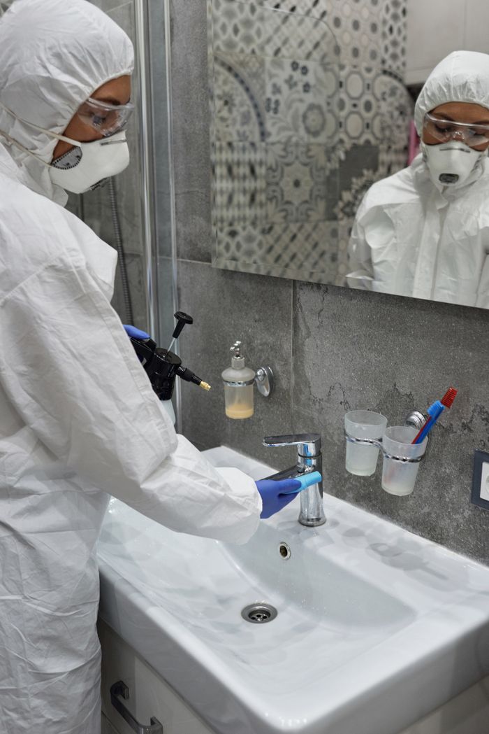 Person in protective gear disinfecting bathroom sink with spray bottle.
